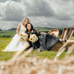 Two brides sitting in front of a tipi at a countryside wedding in Skipton, Yorkshire, holding bouquets and smiling, captured by Yorkshire wedding photographer.