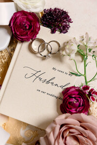 A wedding vow book with the text My vows to my Husband on our wedding day, adorned with a pair of rings and surrounded by flowers, including red and white roses, is placed on a golden surface.