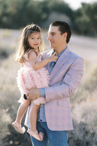 Father holding his young daughter from the Parker family in a soft desert setting in Las Vegas, with the child wearing a pink dress and smiling during a warm golden hour family portrait session.