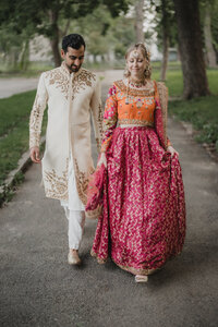 Couple marchant ensemble lors de leur cérémonie du Mehndi, tenues traditionnelles brodées, sherwani crème pour le marié et lehenga rose et orange pour la mariée