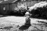 a baby sits in the yard in the sun with a hat on
