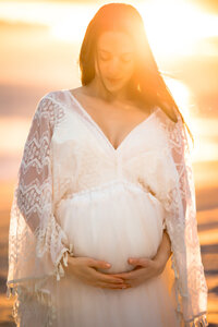 The sun provides a golden backlight to a pregnant mother on the beach. She wears a white dress and looks softly down at her bump while holding it from beneath with both hands.