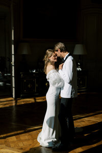Bride and groom standing close together in a softly lit ballroom, sharing an intimate moment as sunlight streams across the wooden floor.