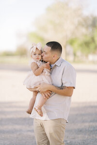 Father gently kisses his baby daughter while holding her during an outdoor family portrait session, captured in soft natural light with a warm, candid moment.