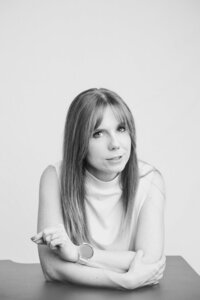 A black and white picture of a blonde woman with straight hair speaking in front of a white backdrop. She wears a white white shirt and a slight smile, while gesturing with one hand. Her arms rest on the black table in front of her.