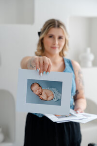 Woman holding a matted newborn portrait print with soft blue tones in a bright studio, Kaitlyn Dawn Photography, Yucaipa newborn photographer.