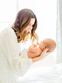 Mother smiling at her newborn baby while holding them in soft window light during an in-home newborn photography session.