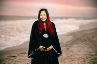 A pregnant woman with black and red hair smiles on a beach. The sun is setting with beautiful pink tones in the sky and she wears a navy blue velvet dress.