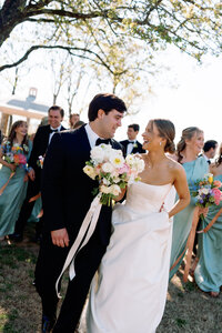 Bride and groom smiling at each other while walking together with bouquet in hand, surrounded by their wedding party at Burritt on the Mountain in Huntsville, Alabama, photographed by Perry Hancock.