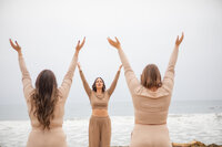 Three women wearing matching neutral-toned outfits raise their arms in a synchronized pose during a mindful movement session on the El Matador beach, with waves crashing behind them.