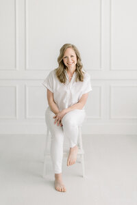 Photo taken in a photography studio of a Dallas TX business woman sitting on a chair while she leans over to pose and smile at the photographer for her professional headshots.