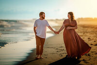 Pregnant couple walking hand in hand along the shoreline during a sunset maternity session in Atlantic Beach North Carolina