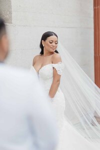 A striking bride poses confidently in an off-the-shoulder lace gown with a long, flowing veil trailing behind her. With soft glam makeup and sleek hair, she stands against a textured stone wall accented by a terracotta column, exuding poise and quiet elegance. The blurred figure of the groom adds depth and movement, making the moment feel spontaneous and cinematic. Captured in Phavy Photography’s signature true-to-color, editorial documentary style—refined, intentional, and full of grace.