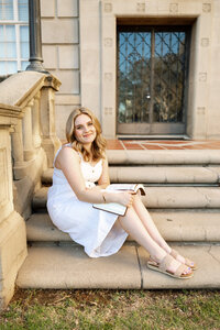 High school senior girl sitting on outdoor stone steps holding a book in a soft natural light setting, senior portrait by Kaitlyn Dawn Photography.