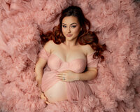 Pregnant woman with red hair lying in layers of a blush pink ruffled maternity gown while gently holding her baby bump during a close-up studio maternity portrait photographed by Inland Empire maternity photographer Kaitlyn Dawn Photography.