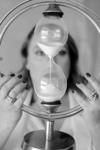 Black and white portrait of a woman viewed through the glass of a large hourglass she holds in both hands.