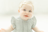 A blue eyed baby girl sitting on the floor in a photography studio for her milestone photos.