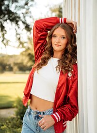 Senior portrait in downtown Magnolia Springs, Alabama — leaning against a white pillar wearing jeans and a red dance team jacket.