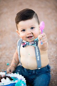 Professional photography by Nature's Child Photography: Toddler boy wearing a bowtie and birthday suspenders with blue frosting on his face, holding a pink spoon during a beach cake smash photography session on Ponte Vedra Beach.