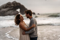 couple embracing at the beach with large rock in the background containing a gap in the shape of a heart