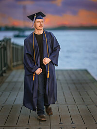 High school senior boy wearing cap and gown on waterfront dock at sunset in New Bern North Carolina