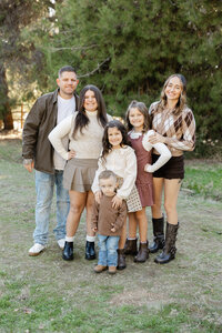 Family of six standing together in a grassy outdoor setting with coordinated earth-tone outfits, joyful candid moment captured by Kaitlyn Dawn Photography, professional family portrait photographer.