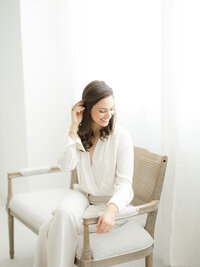 Professional headshot photo of a local Dallas business as she sits on a bench holding her hair away from her face and looking down as she smiles.