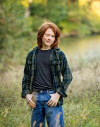 Tween boy with shoulder-length red hair wearing a plaid flannel and jeans standing confidently in a grassy field during an outdoor in Edwardsville, Illinois. (Photographed by Nature's Child Photography)