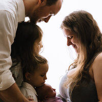 A white baclit picture shows a mother smiling down at her newborn baby while her other two children and husband gaze down at the baby from the other side of the frame.