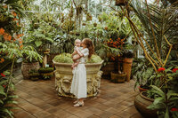Mom in a white dress holding her  baby boy in the middle of beautiful plants at the