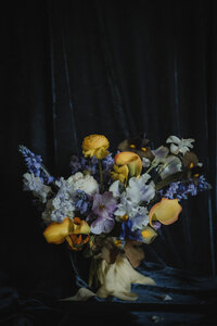Bouquet de mariage composé de renoncules jaunes, delphiniums bleus et roses pâles, éclairage dramatique sur fond de velours sombre, style pictural inspiré des maîtres hollandais