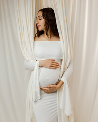 Expecting mom in a white fitted gown gently holding her belly while framed by neutral drapery during a maternity session by Kaitlyn Dawn Photography in Yucaipa.