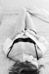 Close up black and white boudoir photo of a woman laying on a bed in a photography studio wearing black lace lingerie looking to her side.