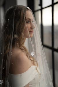 Portrait de mariée de profil sous un voile orné de perles et de fleurs, robe bustier, lumière naturelle près d'une fenêtre au Studio Giovanelli
