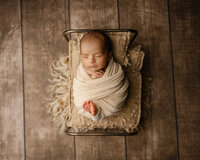 Sleeping newborn baby wrapped in a cream swaddle and posed in a small vintage bed prop on a rustic wooden backdrop during a newborn photography session by Kaitlyn Dawn Photography.