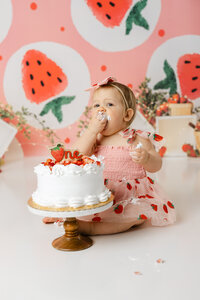 Baby in strawberry dress tasting cake during cake smash session with watermelon backdrop. Photograph by Yucaipa cake smash photographer Kaitlyn Dawn Photography.