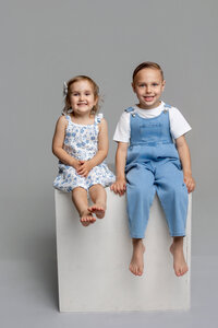 Brother and sister sitting on a white studio cube in a neutral gray studio, coordinated blue outfits and smiling expressions captured by Kaitlyn Dawn Photography, professional children’s and sibling portrait photographer.