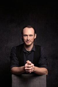 A dark haired man with light blue eyes smiles softly with his hands folded in front of him. He wears a gray shirt with a black vest and sits in front of a gray backdrop. The lighting is dramatic.