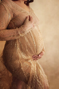 Close-up of a pregnant belly in a sparkling gold gown, hands gently cradling the bump in a glamorous and intimate maternity session near Pewaukee, Wisconsin.