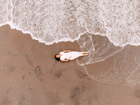 An aerial photograph of a dark haired pregnant woman laying on the beach. The water is cusping the shore and wrapping around her feet.