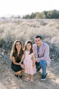 The Parker family sitting together in a soft desert landscape in Las Vegas, with parents and their young daughter smiling at the camera during a warm natural light family portrait session.