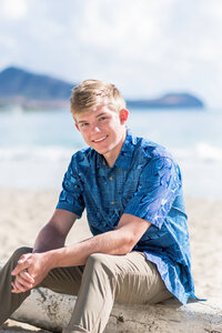 Senior boy sitting on a log on a sandy beach and smiling at the camera.