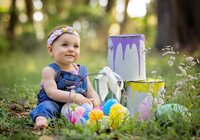 Baby girl in denim overalls and a floral headband sitting in the grass beside painted buckets and colorful Easter eggs during an Easter themed outdoor child portrait session in St. Johns. (Photographed by Nature's Child Photography)