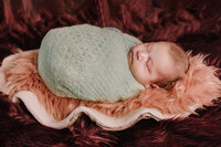 A sleeping baby in a seafoam green wrap lies in a coral fur covered clamshell.