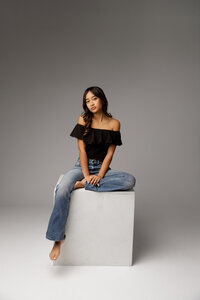 High school senior girl sitting on a white studio block wearing a black top and jeans, studio senior photography by Kaitlyn Dawn Photography.