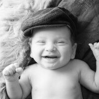 Black and white newborn portrait of a smiling baby wearing a newsboy cap, capturing a joyful expression during a custom newborn photography session.
