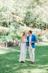 A barefoot bride in a white flowy dress and a groom in a blue blazer share a tender kiss while holding hands in a sunlit garden.
