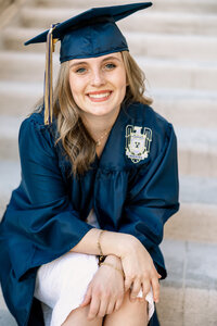 Smiling high school graduate wearing a cap and gown sitting on stone steps during golden hour, graduation portrait by Kaitlyn