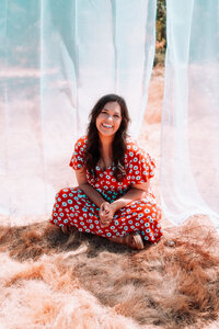 Singer-songwriter smiling outdoors in a red floral dress surrounded by flowing white fabric