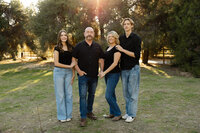 Family of four standing together in a grassy park during golden hour, coordinated casual outfits and warm sunlight captured by Kaitlyn Dawn Photography, professional outdoor family portrait photographer.